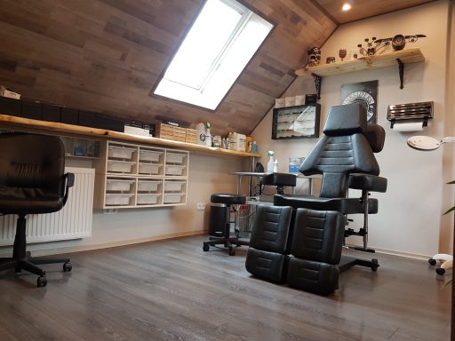 Modernes Tattoo-Studio mit Arbeitstisch, Stuhl und Regalen unter einem Dachfenster.